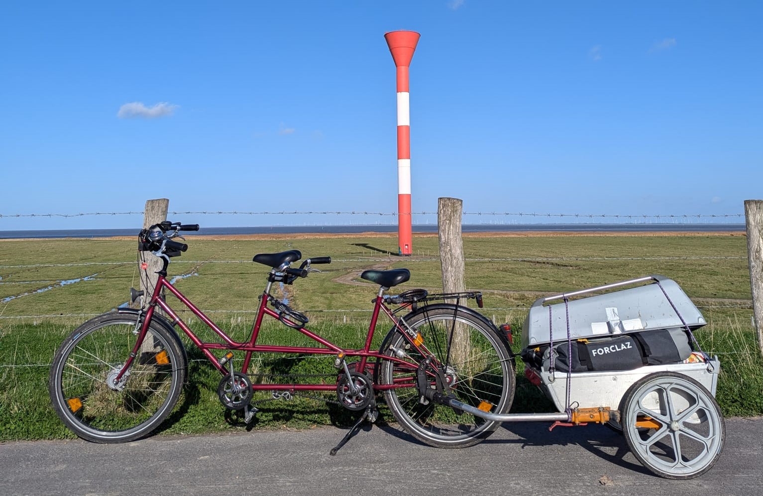 Tandem mit Anhänger vor grüner Weidelandschaft mit rot-weißem Leuchtturm und strahlend blauem Himmel